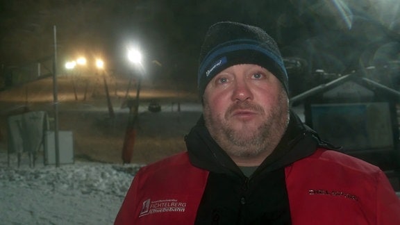Der Geschäftsführer der Fichtelberg-Schwebebahn, René Lötzsch, ein Mann mit Bart und Mütze, steht vor einem Skihang. 