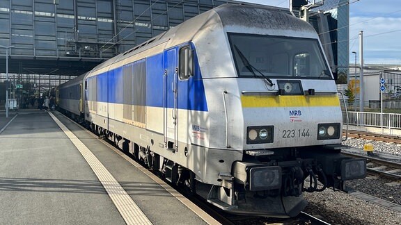 Eine silber-blaue Diesellok steht mit einem Reisezug an einem Bahnsteig