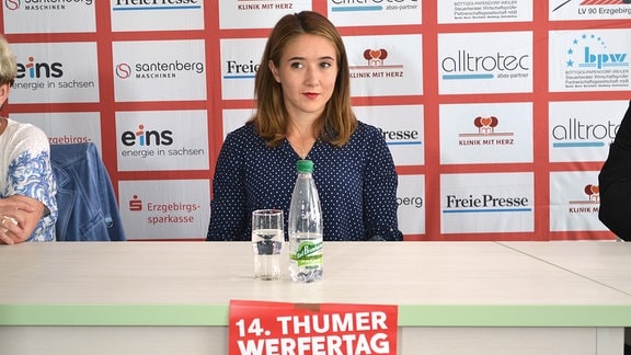 Eine junge Frau sitzt an einem Podium bei einer Pressekonferenz. Im Hintergrund eine Tafel mit Sponsorenlogos, im Vordergrund ist der Auschnitt eines kleinen Plakates mit der Überschrift "Thumer Werfertage" zu sehen. 