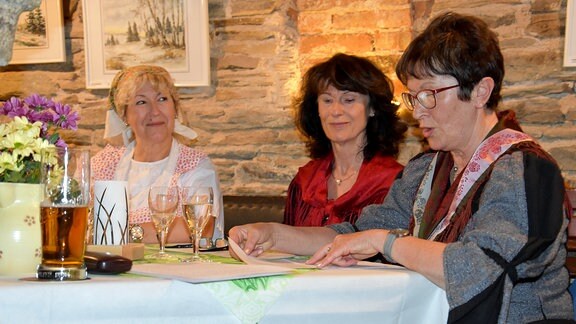 Drei Frauen, eine davon in Tracht, sitzen an einem Tischvor einer Natursteinwand. Eine von ihnen liest einene Text vor
