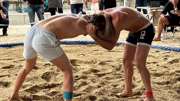 Zwei Männer stehen kämpfend in einer Sand-Arena.