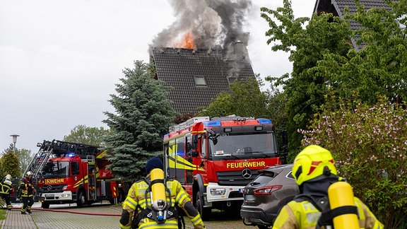 Ein Einfamilienhaus brennt oben im Dach. Feuerwehrleute rennen auf das Haus zu und wollen löschen.