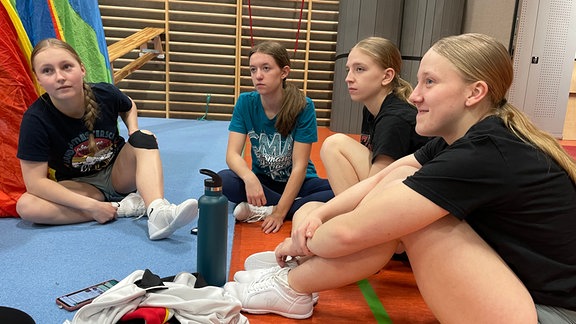 Vier Mädchen in Trainingssachen sitzen auf dem Boden einer Turnhalle.