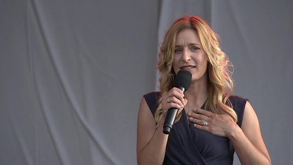 Die Sängerin Stefanie Hertel steht in einem schwarzen, ärmellosen Kleid auf der bühen und spricht zu denBesuchern der Gedenkfeier.