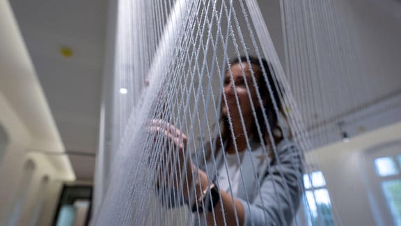 Eine Frau berührt eine Installation aus diagonal übereinander gespannten hellblauen Fäden.
