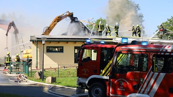 Ein Bagger hebt das Dach eine Gebäudes an, während Feuerwehrleute versuchen den Brand in dem Gebäude zu löschen