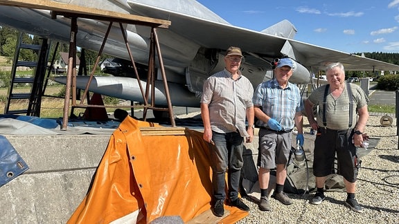 Drei Herren in Arbeitskleidung vor den zu restaurierenden Jagdflugzeug MiG 21