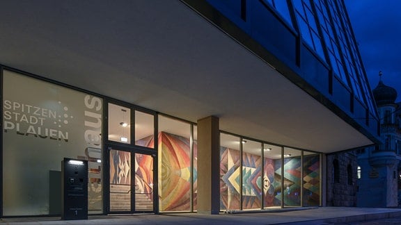 Blick in den verglasten Eingangsbereich eines Gebäudes, in dem ein buntes Wandbild mit geometrischen Formen zu erkennen ist.
