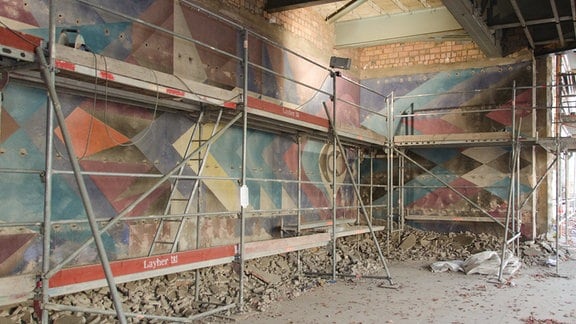 Blick in einen Raum, in dem ein Baugerüst vor einer Wand steht, an der die Reste eines Gemäldes zu sehen sind; auf dem Fußboden liegen Steine und eine Plane