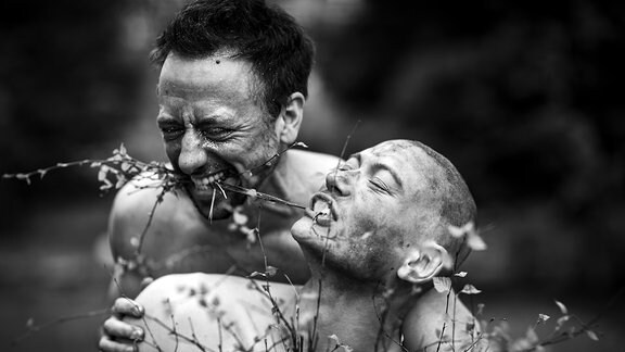 Zwei Personen mit nackten Oberkörpern, einer mit Zweigen im Mund, freuen sich entspannt in der Natur