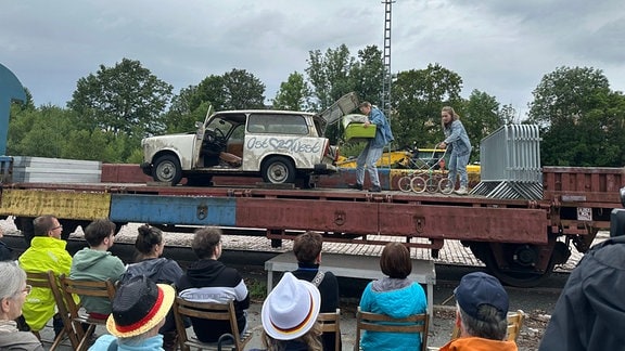 Auf einem offenen Güterzugwagen-Auflieger steht ein Trabi Kombi, in dem von zwei Schauspielern ein Kinderwagen verladen wird. Vor dem Güterwagen sitzen Zuschauer auf Holzstühlen.