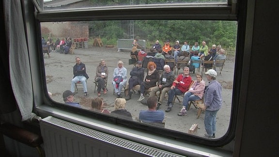 Blick aus einem Zugfenster nach draußen. Es sind mehrere Gruppen von jeweils 16 oval sich gegenübersitzenden Zuschauerstühlen zu sehen. In jeder Gruppe spielt ein Schauspieler oder eine Schauspielerin. 