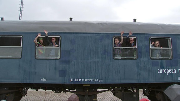 Vier Schauspieler schauen von innen aus zwei Zugfenstern und winken freudestrahlend nach draußen. 