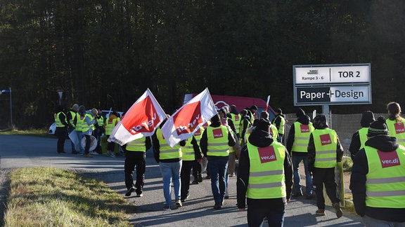 Streikende Mitarbeiter laufen mit Verdi-Fahnen und gelben westen auf ein Werktor zu.  