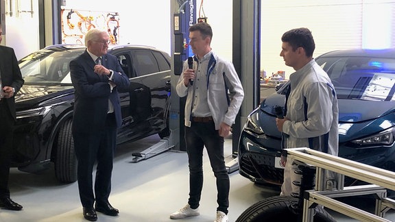 Bundespräsident Frank-Walter Steinmeier steht in einer Werkstatt vor zwei schwarzen Autos und unterhält sich mit zwei Mitarbeitern