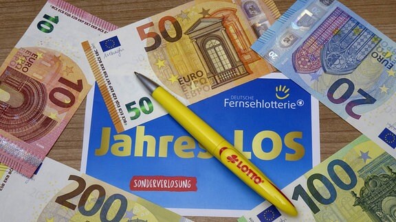 Auf einem Jahreslos der Deutschen Fernsehlotterie liegen Geldscheine und ein Kugelschreiber. 