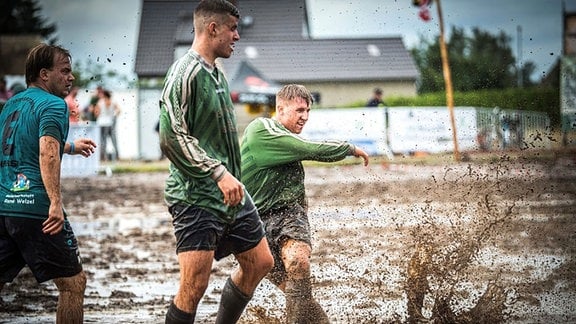 Drei Männer in Fußballtrikots bewegen sich auf einem extrem matschigen Feld. Einer von ihnen hat wohl gerade einen Ball geschossen, der Matsch spritzt meterhoch. 