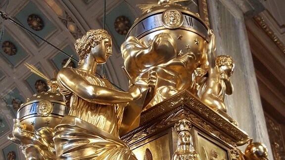 Auf einem Kaminsims in einem Schloss stehen eine große goldene Uhr, davor eine große goldene Figur.
