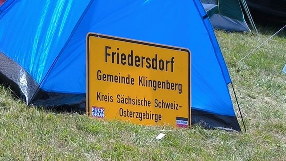 Ein an ein Zelt gelehntes Ortsschild trägt die Aufkleber: "Fuck Grün" und "NS-Zone" auf Reichsflagge.