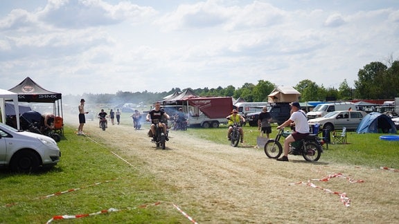 Mehrere junge Männer fahren mit Mopeds auf einem braunen Wiesenpfad. Qualm liegt in der Luft. Drumherum stehen Autos und Zelte.