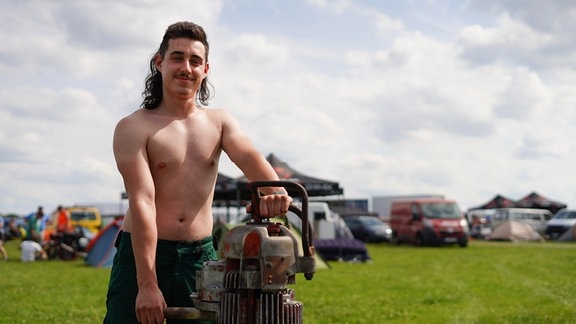 Ein Mann mit Vokuhila steht oberkörperfrei auf einer Wiese. Er hält ein großes Gerät aus Metall fest.