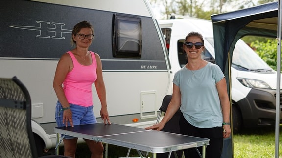 Zwei Frauen stehen an einem Klapptisch vor Wohnwagen. Sie schauen freundlich in die Kamera.