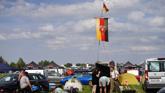Eine DDR-Fahne hängt an einem abgespannten Fahnenmast. Daneben stehen mehrere Menschen. Drumherum sind Zelte und Autos.