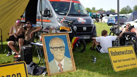 Ein großes Bild von Erich Honecker lehnt an einem Campinglager. In seinem Mund steckt eine Zigarette. Davor stehen zwei gelbe Ortsschilder. Im Hintergrund sitzen Menschen. 