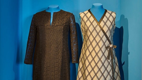 Auf zwei blauen Kleiderständern hängen ein schwarzes und ein weißes Kleid in einer blauen Ausstellungsvitrine.