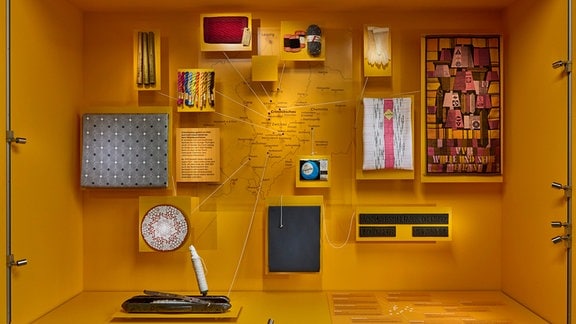 in einer gelben Ausstellungsvitrine sind verschiedene Exponate zu sehen, die durch Fäden miteinander verbunden sind.