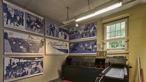 eine alte Maschine aus dunklem Holz ist zu sehen, an der Wand dahinter hängen mehrere in Blautönen gehaltene Bilder im Comicstil.