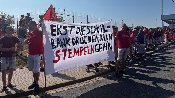 VW-Mitarbeiter in roten T-Shirts der IG Metall stehen vor dem Werksgelände und halten Protestschilder hoch.