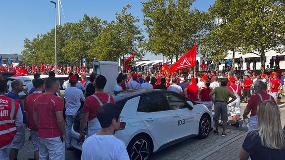 VW-Mitarbeiter in roten T-Shirts der IG Metall stehen vor dem Werksgelände und protestieren.