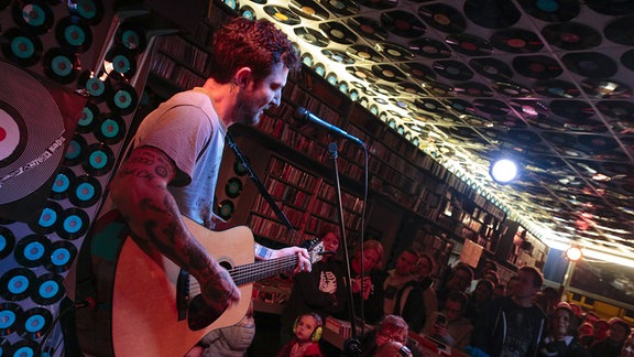 Ein Mann steht mit Gitarre auf einer Bühne vor einem Live-Publikum.