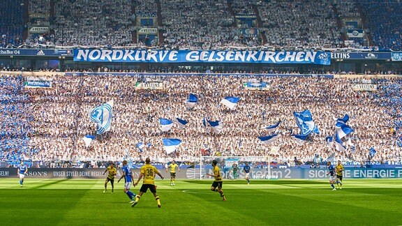 Fußballstadion von Schalke 04 mit Spielern auf dem Spielfeld, vollen Tribünen, Fahnen und dem Spruchband "Nordkurve Gelsenkirchen".