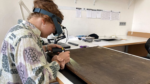 Eine junge Frau mit Schutzbrille sitzt an einem Tisch und beugt sich über ein verschmutztes Ölgemälde.