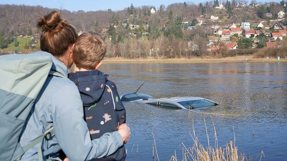 Eine Frau und ein Kind betrachten einen PKW in einem Fluss.