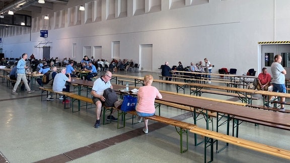 Menschen sitzen an Bänken und Tischen in einer großen Halle.