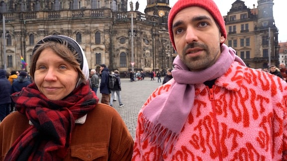 Eine Frau und ein Mann mit Winterkleidung und Schals stehen auf einem Platz. 
