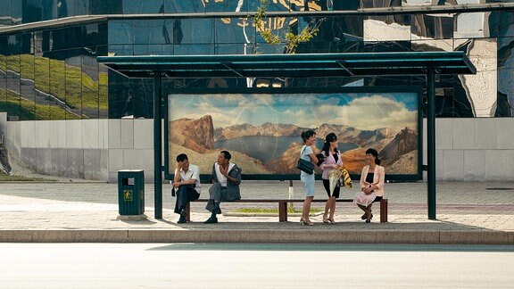 Eine Bushaltestelle in Pjöngjang, an der Menschen sitzen. An der Bushaltestelle ist ein großes Bild von Bergen angebracht.
