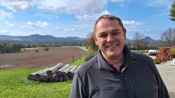 ein Mann mittleren Alters steht auf dem hof eines Grundstücks in der Sächsischen Schweiz und schaut lächelnd in die Kamera.