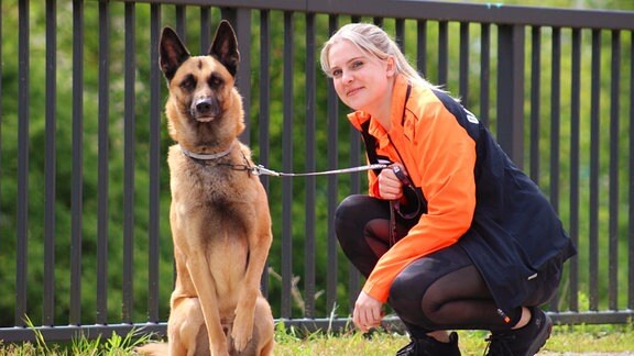 Eine Frau in Sportkleidung hockt neben einem Schäferhund, den sie an der Leine hält.