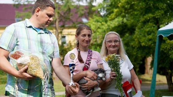 Drei Menschen stehen nebeneinander. Die Frau in der Mitte hält ein Kaninchen. Der Mann und die Frau neben ihr halten Blumen, Hasenfutter und einen Blumenstrauß.