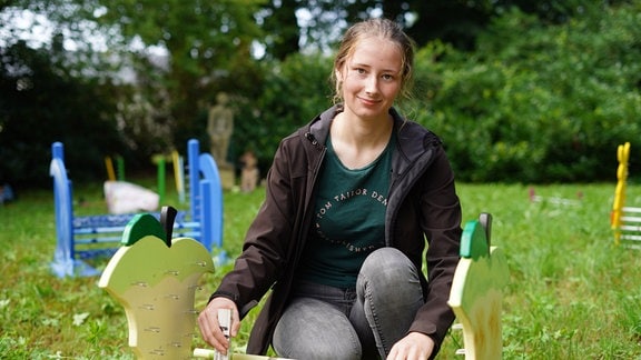 Eine junge Frau kniet auf einer Wiese an einer Hürde und schaut in die Kamera.