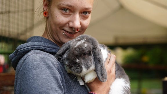 Eine Frau hält ein Kaninchen nah an ihrer Brust und schaut in die Kamera.