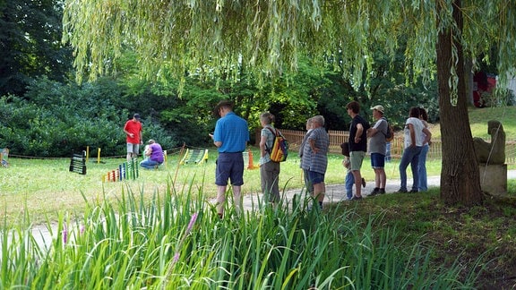 Eine Reihe von Menschen stehen auf einem Weg in einem Park und schauen auf eine Wiese mit einem Hindernissparcours.