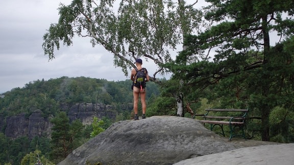 Ein nackter Mann steht mit dem Rücken zur Kamera auf einem Felsen, in einer bergigen Gegend.