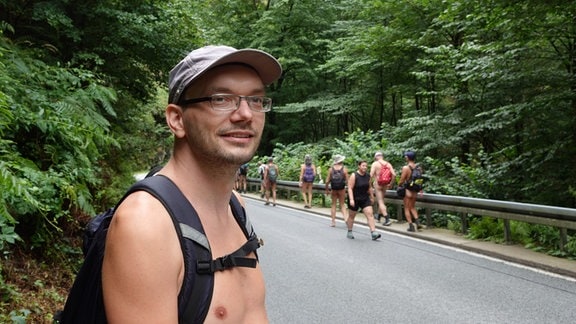 Im Vordergrund steht ein Man mit Brille und Basecap. Im Hintergrund läuft eine Gruppe menschen an einer Straße entlang. Fast alle sind unbekleidet.