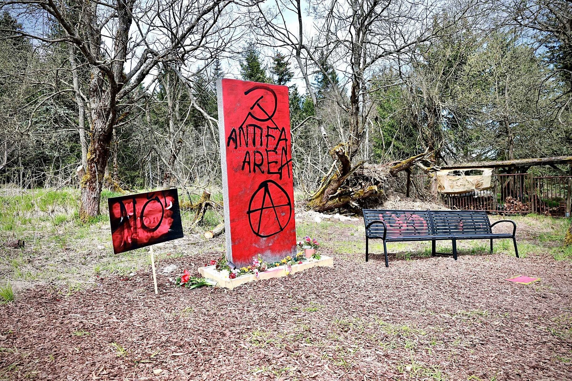 Auf einer Wiese in Zinnwald stehen ein Schild, ein Gedenkstein und eine Bank, die mit roter und schwarzer Farbe besprüht sind