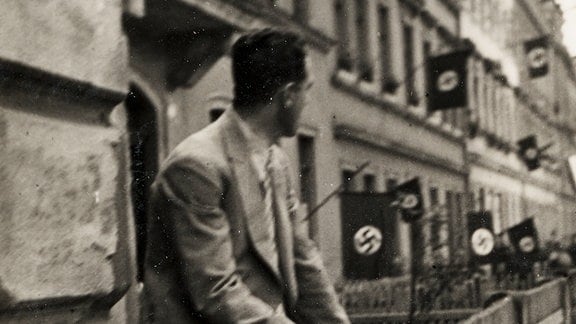 Schwarzweißaufnahme eines Mann, der in den 1930er-Jahren in Dresden auf der Mauer vor einer Häuserzeile sitzt, an deren Fassade Hakenkreuzfahnen wehen 